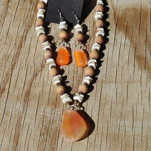 May include: A handmade jewelry set featuring a necklace and matching earrings. The necklace and earrings are made with wooden beads, white shell accents, and orange-colored glass pendants. The jewelry is displayed on a wooden surface.