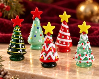 Peut inclure: Cinq figurines de sapins de Noël en verre miniature, chacune surmontée d'une étoile. Les arbres présentent divers motifs, notamment vert uni avec des lignes jaunes, vert clair avec des points, rouge avec des zigzags blancs et rouge avec des rayures blanches.