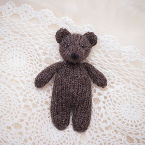 May include: A small, handmade brown knitted teddy bear with black eyes and a black nose. The bear is standing on a white, lacy, crocheted doily. The background is a soft, neutral color.
