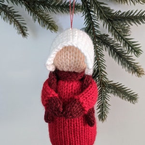 May include: A knitted Christmas ornament shaped like a person wearing a red sweater, white hat, and mittens.