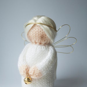 Puede incluir: Muñeca de ángel tejida a mano con vestido blanco, cabello rubio y halo y alas doradas. El ángel sostiene una pequeña campana dorada. Un objeto decorativo suave.