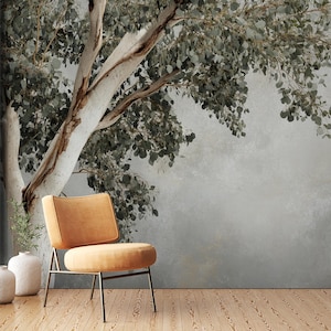Puede incluir: Un sillón amarillo mostaza con estructura de metal se encuentra frente a un mural de pared que presenta un árbol grande de tronco blanco con hojas verdes. El fondo es gris claro con un efecto texturizado sutil.