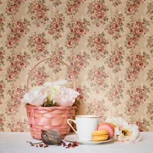 May include: A pink woven basket filled with white and pink flowers sits next to a white teacup and saucer with gold trim. Two macarons and a tea infuser are also present. The background features a floral wallpaper with pink roses.