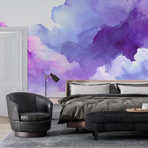 May include: A bedroom with a vibrant purple and blue watercolor mural on the wall. The room features a bed with white pillows, a black armchair, a black ottoman, and a wooden door. A black lamp is on the nightstand.