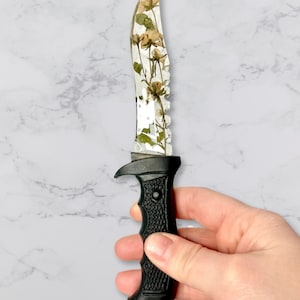 Op de afbeelding: Een mes met een zwart handvat en een doorzichtige kling met daarin ingebedde gedroogde witte bloemen.