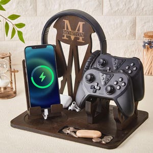 Könnte beinhalten: Gaming-Station aus Holz mit Ladetelefon, Kopfhörern und zwei Gamecontrollern. Das Telefon zeigt ein Ladesymbol an. Die Station ist mit dem Namen "MORTON" graviert.
