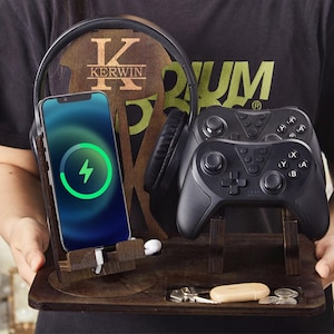 Peut inclure: Station de charge en bois avec un téléphone, un casque et deux manettes de jeu noires. Le téléphone affiche un symbole de chargement. La station a un compartiment pour les clés et les petits objets. Le casque a un accent en bois avec le nom "KERWIN".