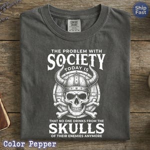 Puede incluir: Camiseta gris oscuro con cuello redondo. La camiseta presenta un gr&aacute;fico blanco de una calavera con un casco con cuernos, con el texto "THE PROBLEM WITH SOCIETY TODAY IS THAT NO ONE DRINKS FROM THE SKULLS OF THEIR ENEMIES ANYMORE."