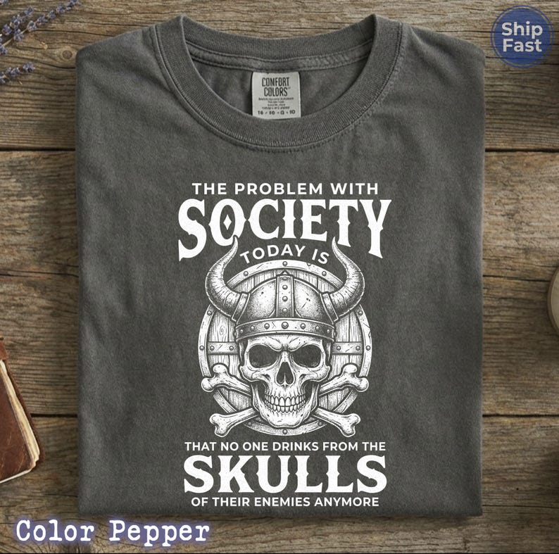Puede incluir: Camiseta gris oscuro con cuello redondo. La camiseta presenta un gr&aacute;fico blanco de una calavera con un casco con cuernos, con el texto "THE PROBLEM WITH SOCIETY TODAY IS THAT NO ONE DRINKS FROM THE SKULLS OF THEIR ENEMIES ANYMORE."