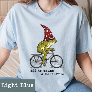 May include: Light blue t-shirt featuring a whimsical illustration of a green frog wearing a red and white polka-dot hat, riding a bicycle. The text "off to cause a kerfuffle" is below the image. The shirt is light blue.