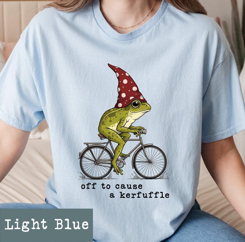 May include: Light blue t-shirt featuring a whimsical illustration of a green frog wearing a red and white polka-dot hat, riding a bicycle. The text "off to cause a kerfuffle" is below the image. The shirt is light blue.