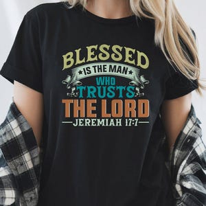 May include: Black t-shirt featuring the quote "BLESSED IS THE MAN WHO TRUSTS THE LORD - JEREMIAH 17:7" in yellow, teal, and orange. The design includes stars and decorative elements. The shirt has a crew neck.