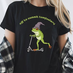 May include: Black t-shirt featuring a green frog wearing a wizard hat and riding a scooter. The text "off to commit tomfoolery" is arched above the frog in green. The frog has orange eyes and feet.