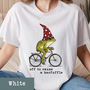 May include: White t-shirt featuring a whimsical illustration of a green frog riding a bicycle and wearing a red mushroom cap with white polka dots. The text "off to cause a kerfuffle" is printed below the image.