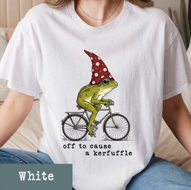 May include: White t-shirt featuring a whimsical illustration of a green frog riding a bicycle and wearing a red mushroom cap with white polka dots. The text "off to cause a kerfuffle" is printed below the image.