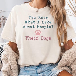 以下が含まれることがあります： 「You Know What I Like About People? Their Dogs」とターコイズとピンクの文字がプリントされたクリーム色のTシャツ。ピンクの足跡のグラフィックがテキストの下に配置されています。カジュアルなスタイルのシャツです。