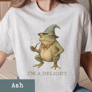 May include: Light grey t-shirt featuring a whimsical illustration of a frog wearing a hat, vest, and bow tie, smoking a pipe. The text "I'M A DELIGHT" is printed below the frog. The shirt is made of a soft material.
