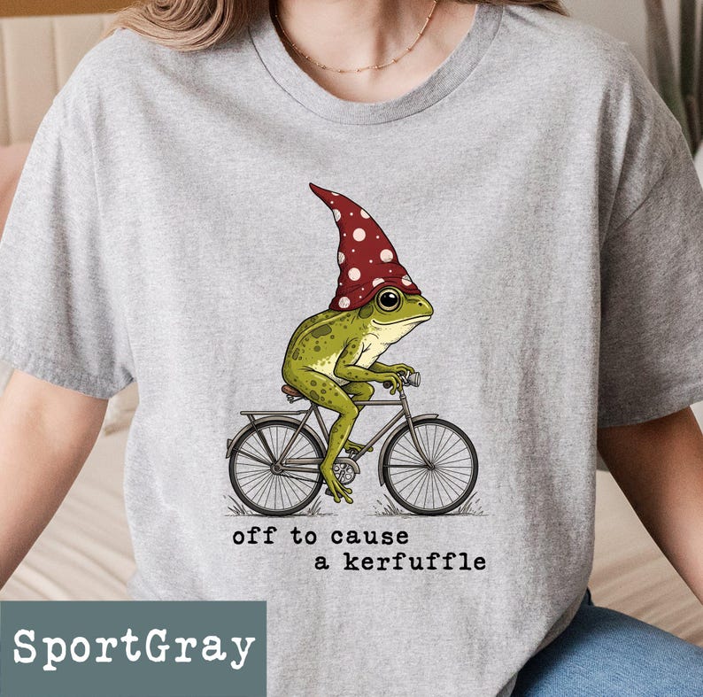 May include: SportGray t-shirt featuring a whimsical illustration of a green frog wearing a red mushroom cap, riding a bicycle. The text "off to cause a kerfuffle" is printed below the image. The shirt is a light gray color.