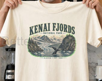 Könnte beinhalten: Ein cremefarbenes T-Shirt mit einer Grafik des Kenai Fjords Nationalparks. Das Design zeigt eine Walflosse im Wasser, schneebedeckte Berge, Gletscher und Tannen, ergänzt durch den Text "KENAI FJORDS NATIONAL PARK ALASKA EST. 1980".