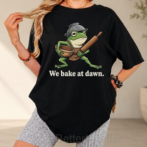 May include: A black t-shirt featuring a graphic of a green frog wearing a colander as a hat, holding a wooden rolling pin and a mixing bowl. The text "We bake at dawn." is printed in white below the frog illustration on the front of the shirt.