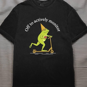 Könnte beinhalten: Schwarzes T-Shirt mit einer Cartoon-Fröschin auf einem Roller, die einen Kegelhut trägt und einen Apfel hält. Der Text "Off to actively monitor" ist über der Fröschin angeordnet. Das Design verwendet Gelb, Grün und Rot.