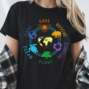 Op de afbeelding: Zwart T-shirt met een kleurrijk ontwerp over milieubescherming. Het ontwerp bevat een bij, neushoorn, schildpad en boom, met de woorden "Save, Rescue, Recycle, Clean, Care, Plant" rond een hartvormige aarde.