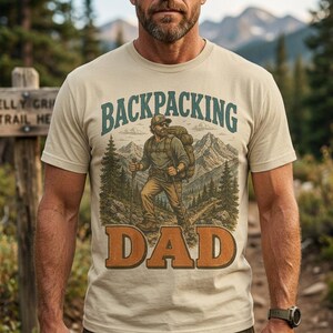 Può includere: Una t-shirt beige a maniche corte con un'illustrazione grafica di un escursionista con zaino e bastoncini da trekking in un paesaggio montano. Il testo BACKPACKING è stampato in verde acqua sopra l'escursionista e DAD in arancione sotto.