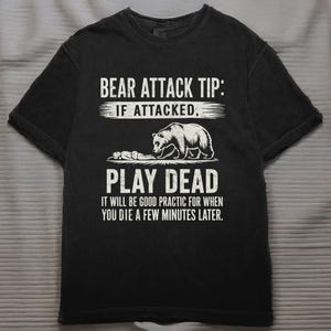 Puede incluir: Camiseta negra con texto y gráfico en blanco. El texto dice: "CONSEJO DE ATAQUE DE OSO: SI ES ATACADO, HAZTE EL MUERTO. SERÁ UNA BUENA PRÁCTICA PARA CUANDO MUERAS UNOS MINUTOS DESPUÉS." Se ilustra un oso y una persona.