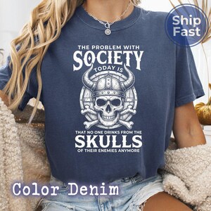 Puede incluir: Una camiseta azul marino con un gr&aacute;fico que presenta una calavera con un casco con cuernos, huesos cruzados y el texto "THE PROBLEM WITH SOCIETY TODAY IS THAT NO ONE DRINKS FROM THE SKULLS OF THEIR ENEMIES ANYMORE". La camiseta est&aacute; etiquetada como "Color Denim".