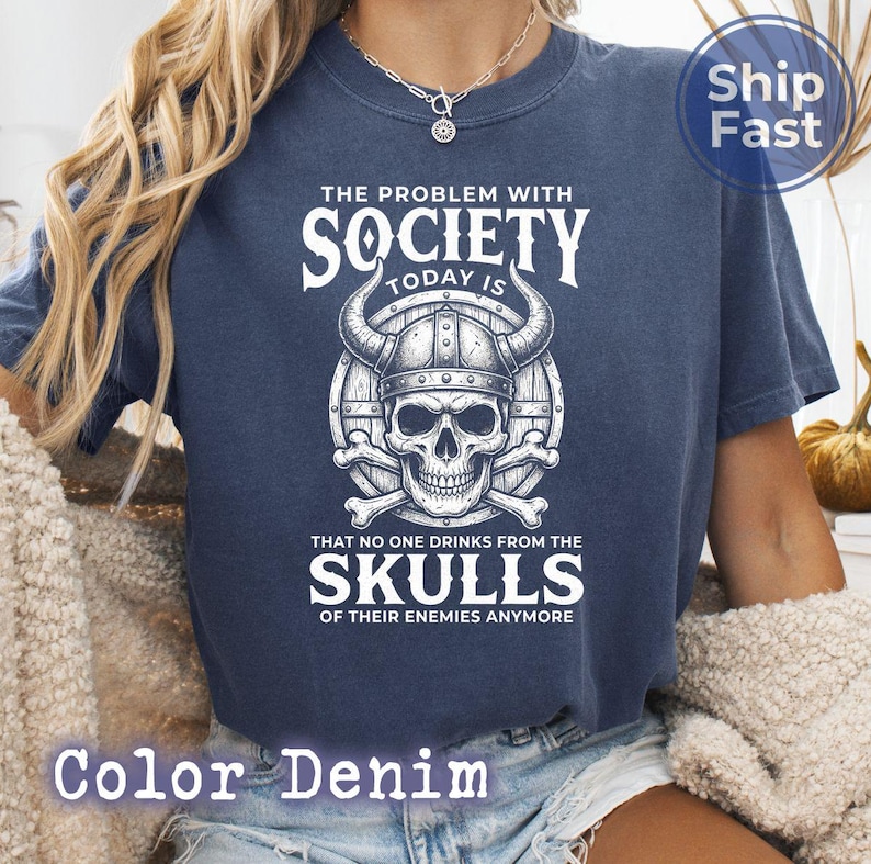 Puede incluir: Una camiseta azul marino con un gr&aacute;fico que presenta una calavera con un casco con cuernos, huesos cruzados y el texto "THE PROBLEM WITH SOCIETY TODAY IS THAT NO ONE DRINKS FROM THE SKULLS OF THEIR ENEMIES ANYMORE". La camiseta est&aacute; etiquetada como "Color Denim".