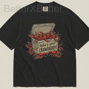 Può includere: T-shirt nero con una grafica di una ghiacciaia piena di gamberi rossi. La ghiacciaia ha la scritta "Who's your CRAWADDY!". I gamberi sono di un rosso vibrante e la maglietta ha un aspetto vintage.