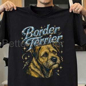 May include: A black short-sleeved t-shirt featuring a graphic of a brown Border Terrier dog. Above the dog, the text Border Terrier is printed in a metallic blue and silver font. The design includes small yellow stars and lightning bolt accents.