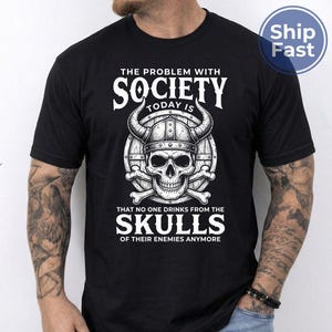 Puede incluir: Camiseta negra con un gr&aacute;fico blanco que presenta una calavera con un casco con cuernos, huesos cruzados y el texto "THE PROBLEM WITH SOCIETY TODAY IS THAT NO ONE DRINKS FROM THE SKULLS OF THEIR ENEMIES ANYMORE."