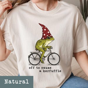 May include: A Natural-colored t-shirt features a whimsical illustration of a green frog wearing a red and white polka-dotted hat, riding a bicycle. The text "off to cause a kerfuffle" is printed below the image.