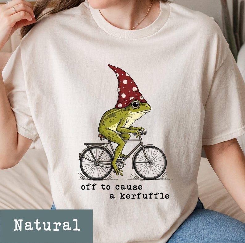 May include: A Natural-colored t-shirt features a whimsical illustration of a green frog wearing a red and white polka-dotted hat, riding a bicycle. The text "off to cause a kerfuffle" is printed below the image.