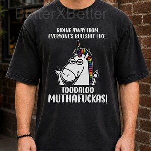 May include: Black t-shirt with a unicorn graphic giving the middle finger. The text reads "RIDING AWAY FROM EVERYONE'S BULLSHIT LIKE... TOODALOO MUTHAFUCKAS!" The unicorn has a rainbow mane and horn. The shirt has a vintage, worn look.