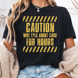 Puede incluir: Camiseta negra con texto amarillo y rayas de peligro. El texto dice "CAUTION WILL TALK ABOUT CARS FOR HOURS". La camiseta es de cuello redondo y tiene mangas cortas. El texto es en negrita, sin serifa.