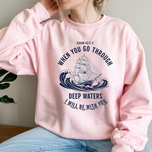 May include: A light pink sweatshirt featuring a navy blue illustration of a sailboat on waves. The text reads "WHEN YOU GO THROUGH DEEP WATERS I WILL BE WITH YOU" and "ISAIAH 43:2-3".