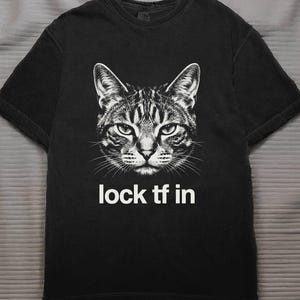 May include: Black t-shirt featuring a detailed grayscale illustration of a cat's face. Below the cat image, the text "lock tf in" is printed in white. The shirt has short sleeves and a crew neck.
