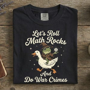 May include: Black t-shirt with the slogan "Let's Roll Math Rocks And Do War Crimes." The design shows a frog in a wizard hat riding a white duck and holding a die.