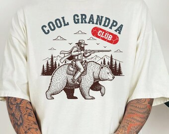 May include: Cream-colored t-shirt with a graphic of a man riding a bear and holding a rifle. The text "COOL GRANDPA CLUB" is above the image in blue and red. The design includes trees, clouds, and birds.