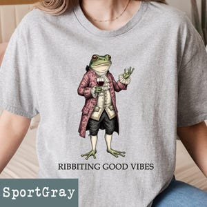 May include: SportGray t-shirt featuring a whimsical illustration of a frog dressed in a vintage coat, holding a wine glass. The frog illustration is accompanied by the text "RIBBITING GOOD VIBES". The shirt is a light gray color.