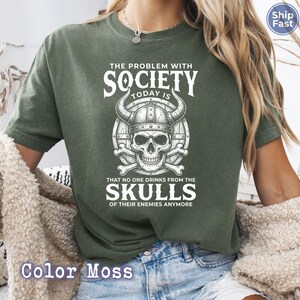 Puede incluir: Una camiseta verde musgo con un gr&aacute;fico que presenta una calavera con un casco con cuernos y el texto "THE PROBLEM WITH SOCIETY TODAY IS THAT NO ONE DRINKS FROM THE SKULLS OF THEIR ENEMIES ANYMORE".