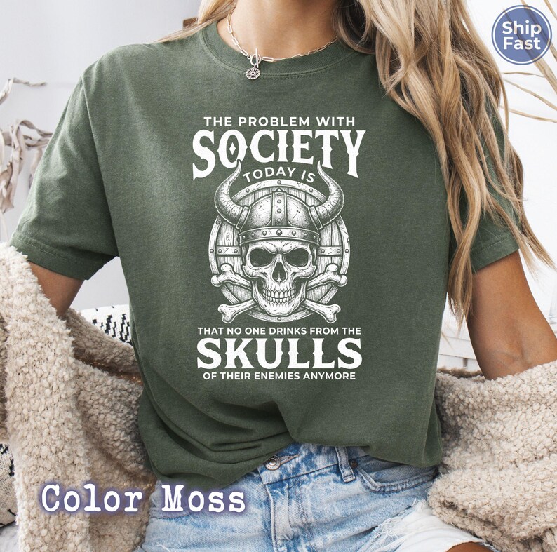 Puede incluir: Una camiseta verde musgo con un gr&aacute;fico que presenta una calavera con un casco con cuernos y el texto "THE PROBLEM WITH SOCIETY TODAY IS THAT NO ONE DRINKS FROM THE SKULLS OF THEIR ENEMIES ANYMORE".
