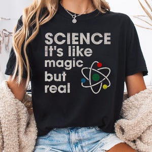 Peut inclure: T-shirt noir avec un graphique blanc de style vintage qui dit "SCIENCE C'est comme de la magie mais réel" et un atome stylisé avec des points colorés.