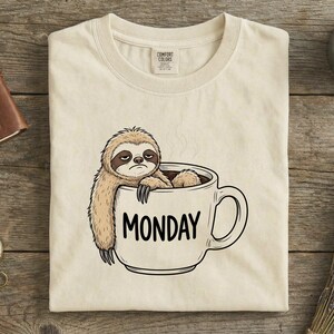 May include: Cream-colored t-shirt featuring a sloth in a coffee mug with the word "MONDAY" in black. The sloth has a grumpy expression. The shirt has a Comfort Colors tag.