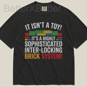 May include: Dark gray t-shirt with the text "IT ISN'T A TOY! IT'S A HIGHLY SOPHISTICATED INTER-LOCKING BRICK SYSTEM!" in white, red, yellow, and blue. The design includes colorful brick illustrations.