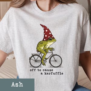 May include: Ash-colored t-shirt featuring a whimsical illustration of a green frog wearing a red and white polka-dot hat, riding a bicycle. The text "off to cause a kerfuffle" is printed below the graphic.