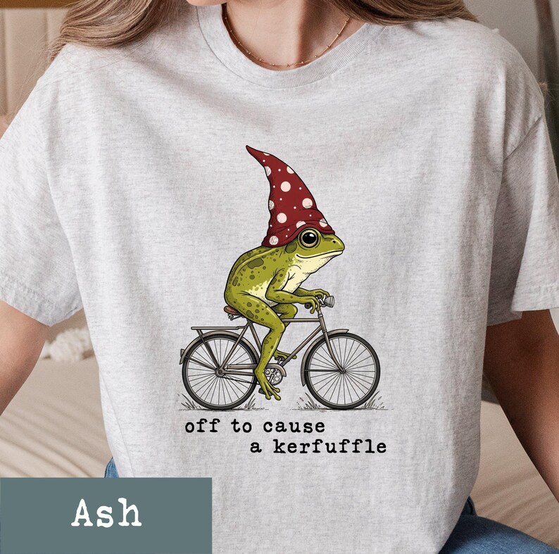 May include: Ash-colored t-shirt featuring a whimsical illustration of a green frog wearing a red and white polka-dot hat, riding a bicycle. The text "off to cause a kerfuffle" is printed below the graphic.