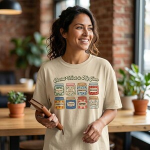 May include: A beige t-shirt featuring a graphic of eight colorful jars, each labeled with social work themes including Empathy, Cultural Awareness, Organization, Advocacy, Administration, Psychosocial Support, and Active Listener, with the text Social Work is My Jam.